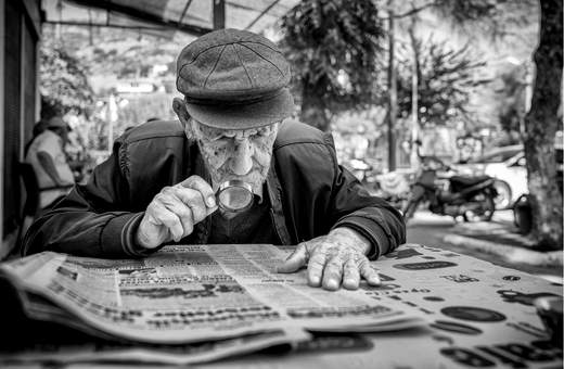 Oudere man met pet leest de krant met een vergrootglas